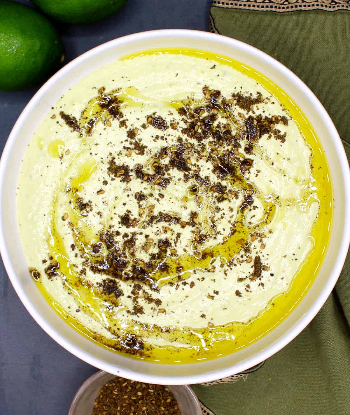 A bowl of fluffy, creamy edamame hummus with za'atar and olive oil. Limes are in the background.