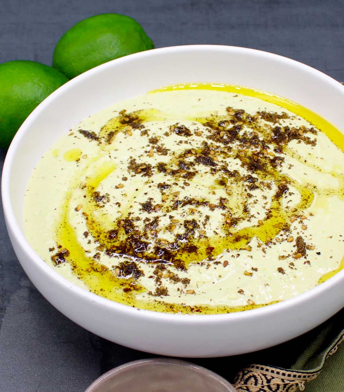 Edamame-Humus in einer weißen Schüssel mit Olivenöl und Za'atar und Geren-Serviette auf der Seite.  Es gibt Zitronen im Hintergrund.