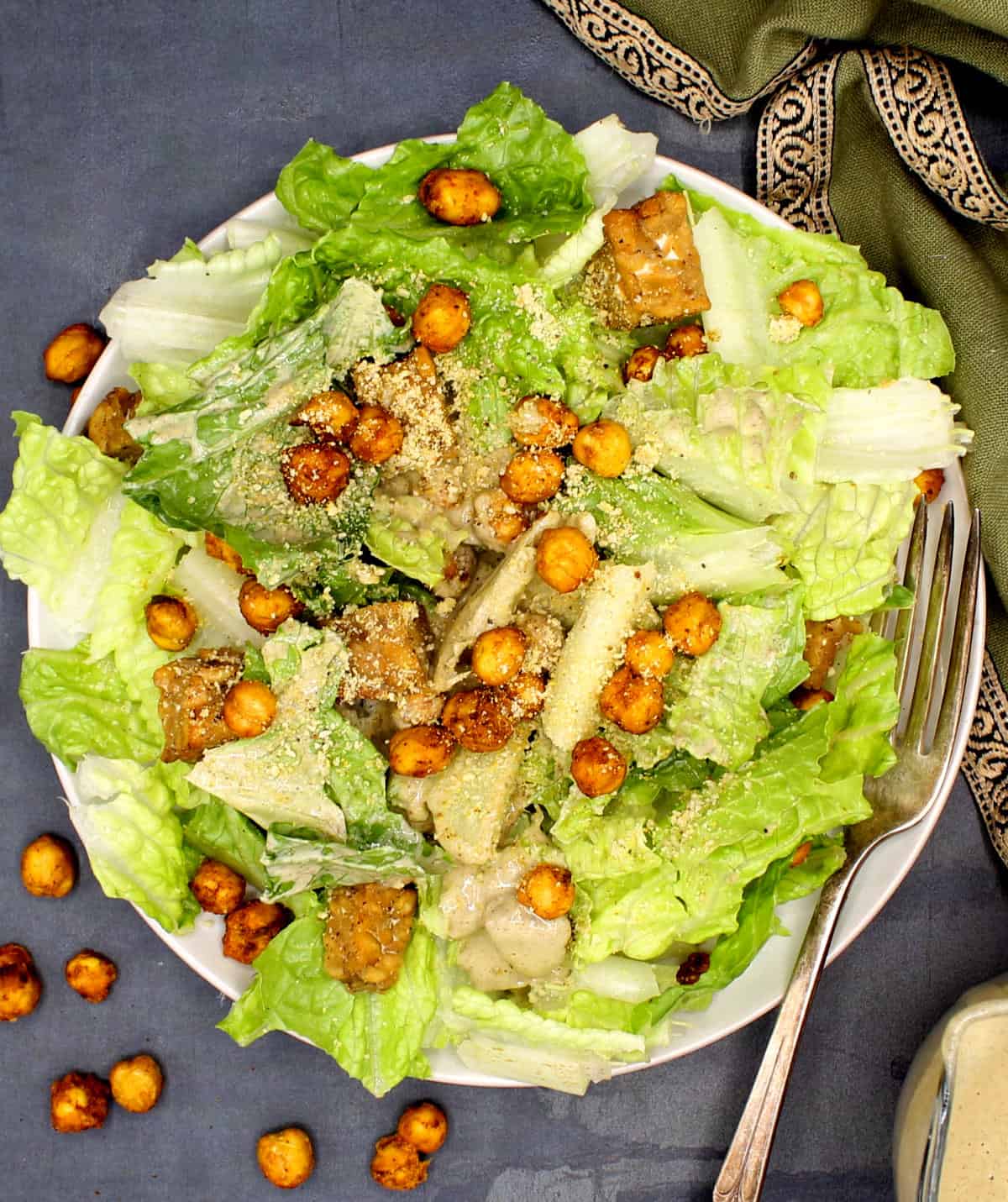 A white plate with vegan Caesar salad made with romaine lettuce, chickpeas, tempeh and vegan parmesan. Chickpeas are scattered on the side,