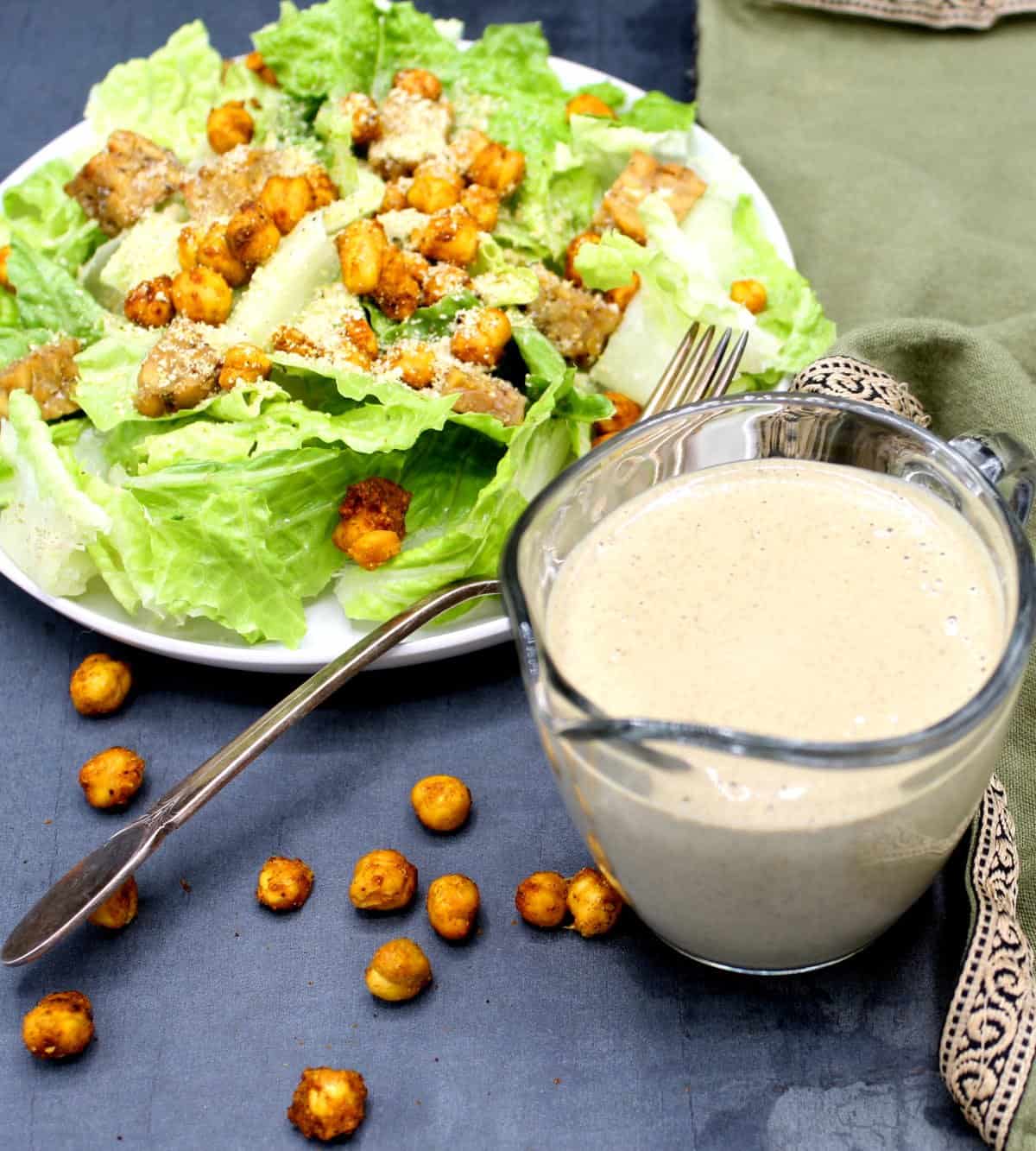 A glass beaker with vegan caesar salad dressing and a plate of vegan caesar salad with tempeh and chickpea croutons.