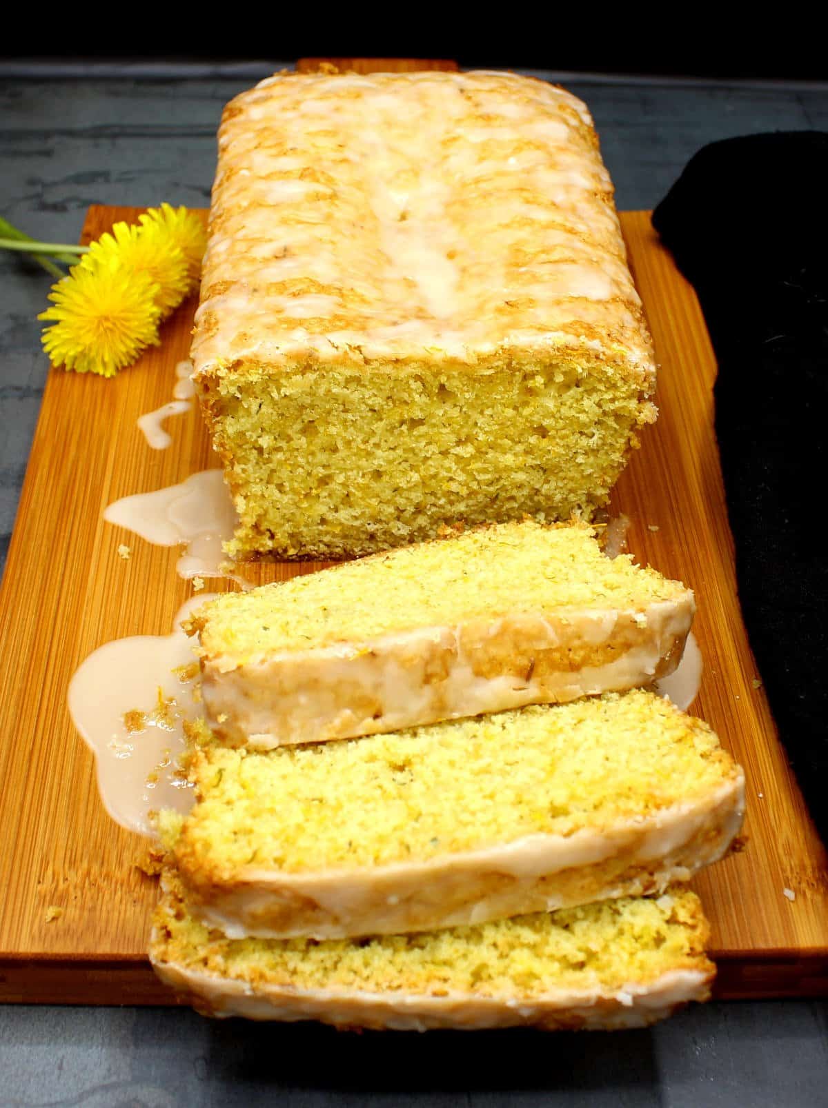 Sliced dandelion bread with lemon glaze on a chopping board with dandelion blossoms on the side.