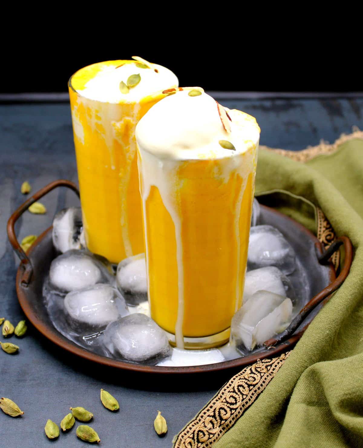 Two glasses of vegan mango smoothie topped with vegan ice cream and an almond and pumpkin seed garnish. The glasses are standing in a tray with cubes of ice and cardamom pods are strewn around.