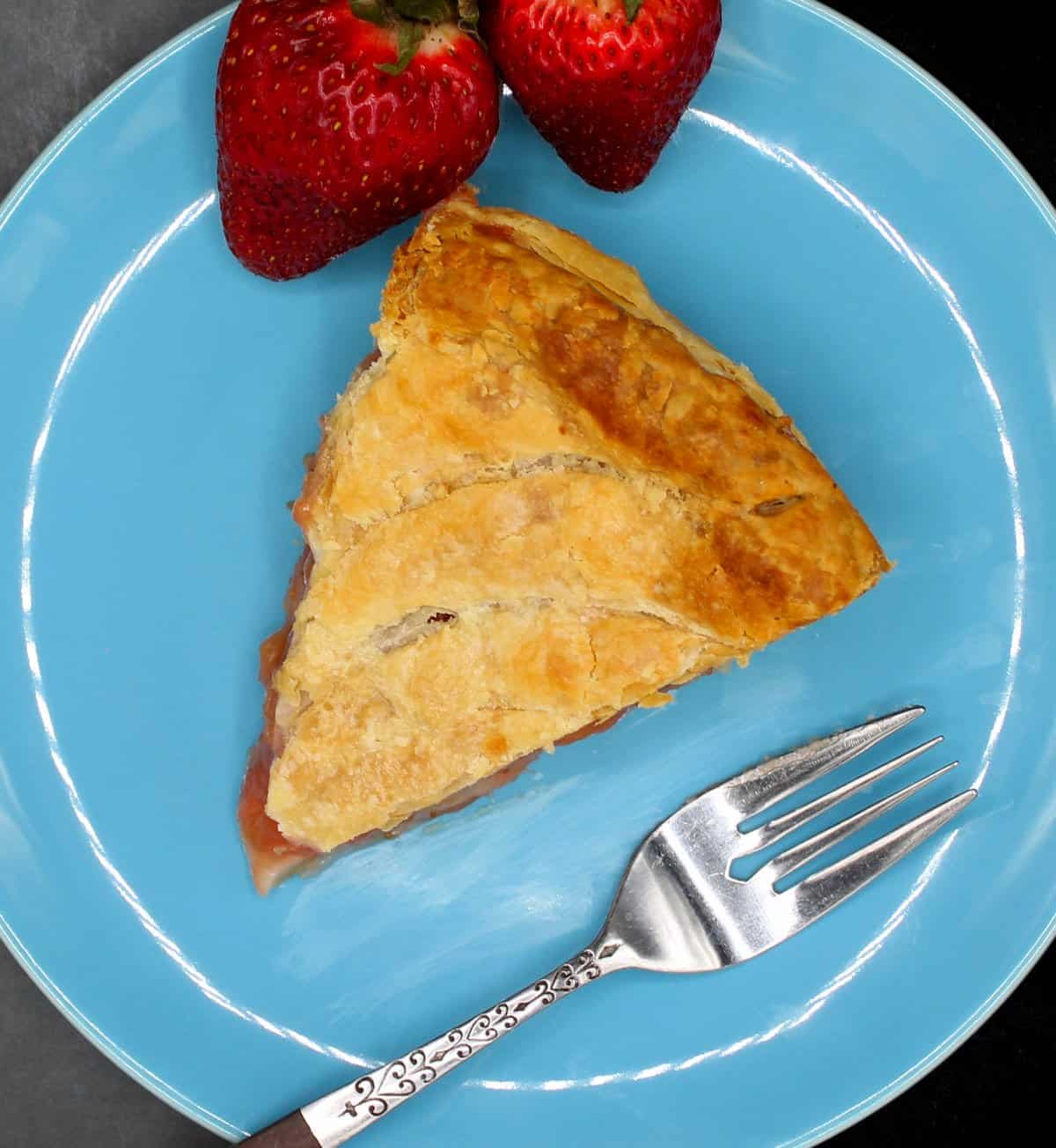 A slice of vegan strawberry rhubarb pithiviers in a blue plate with fork abd two strawberries.