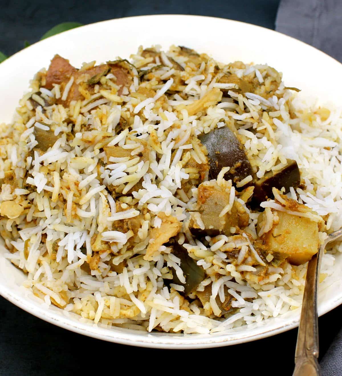 A white bowl with spicy eggplant biryani with chunks of brinjal and potato and a fork.