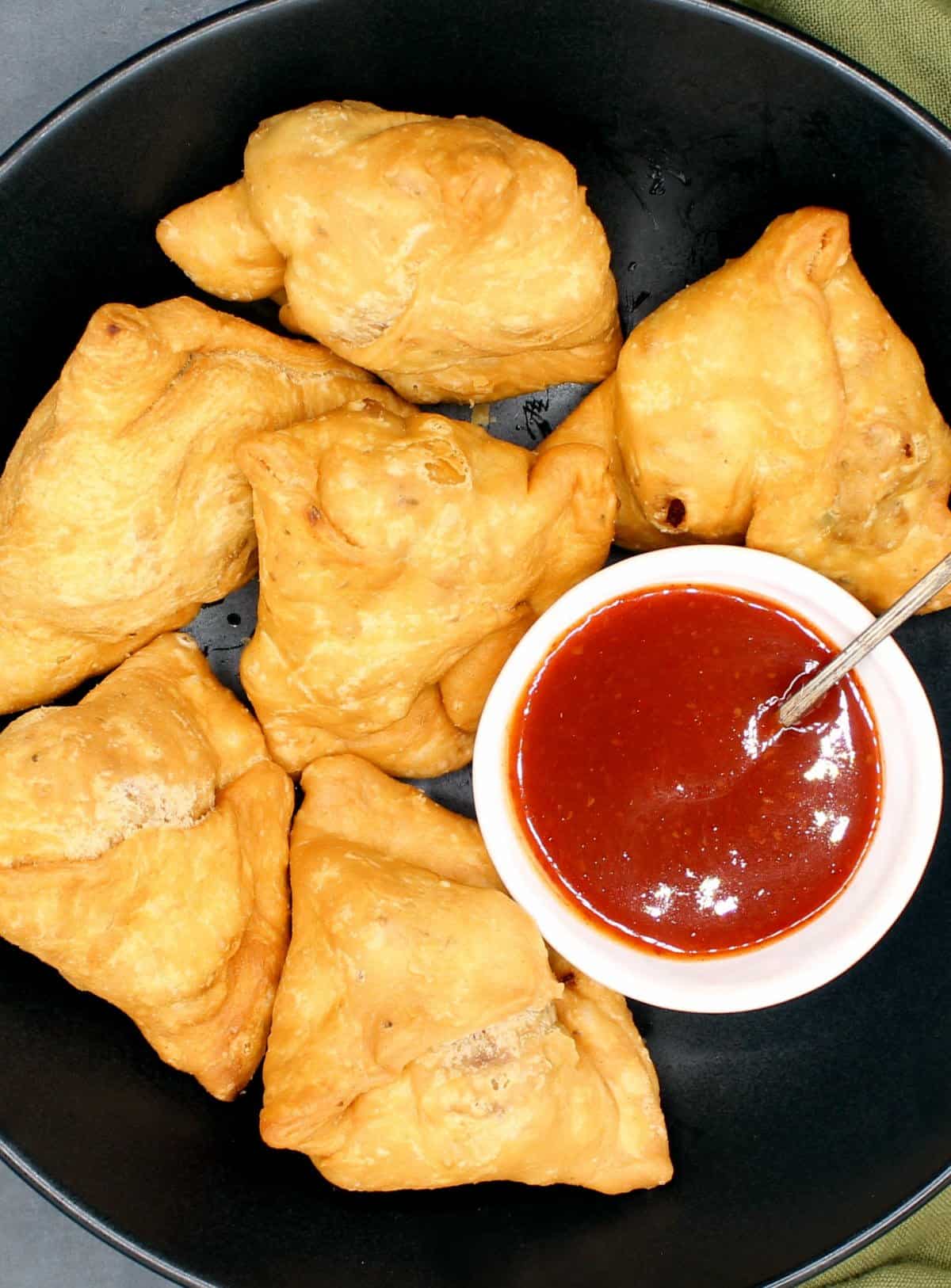 Six golden samosas on a black plate with ketchup in a bowl.