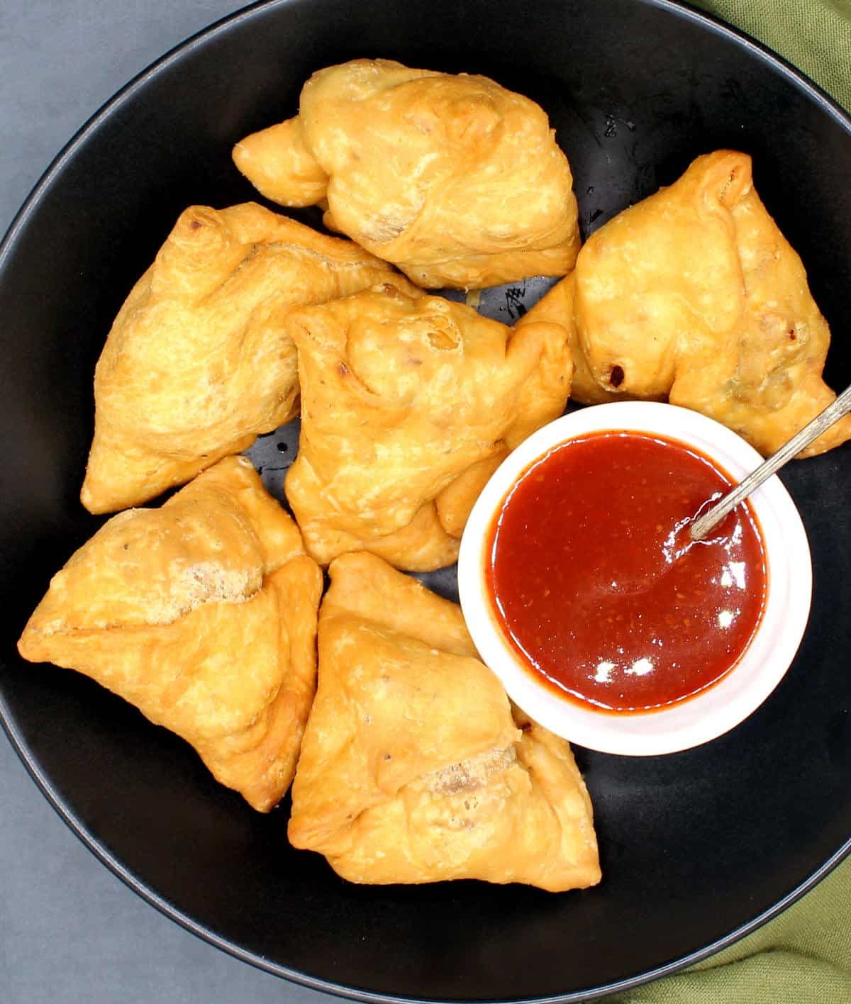 A black bowl with six samosas and a red sauce for dipping.