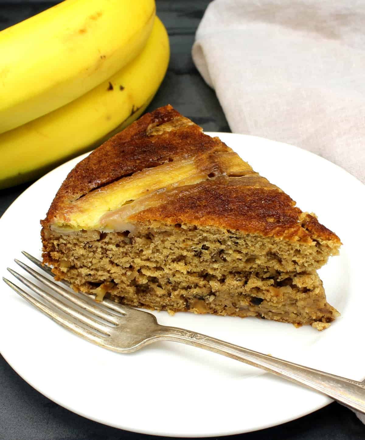 Slice of vegan banana cake with a fork and bananas in background.