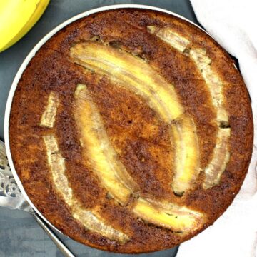 Vegan banana cake on white plate with bananas and cake server partially visible.