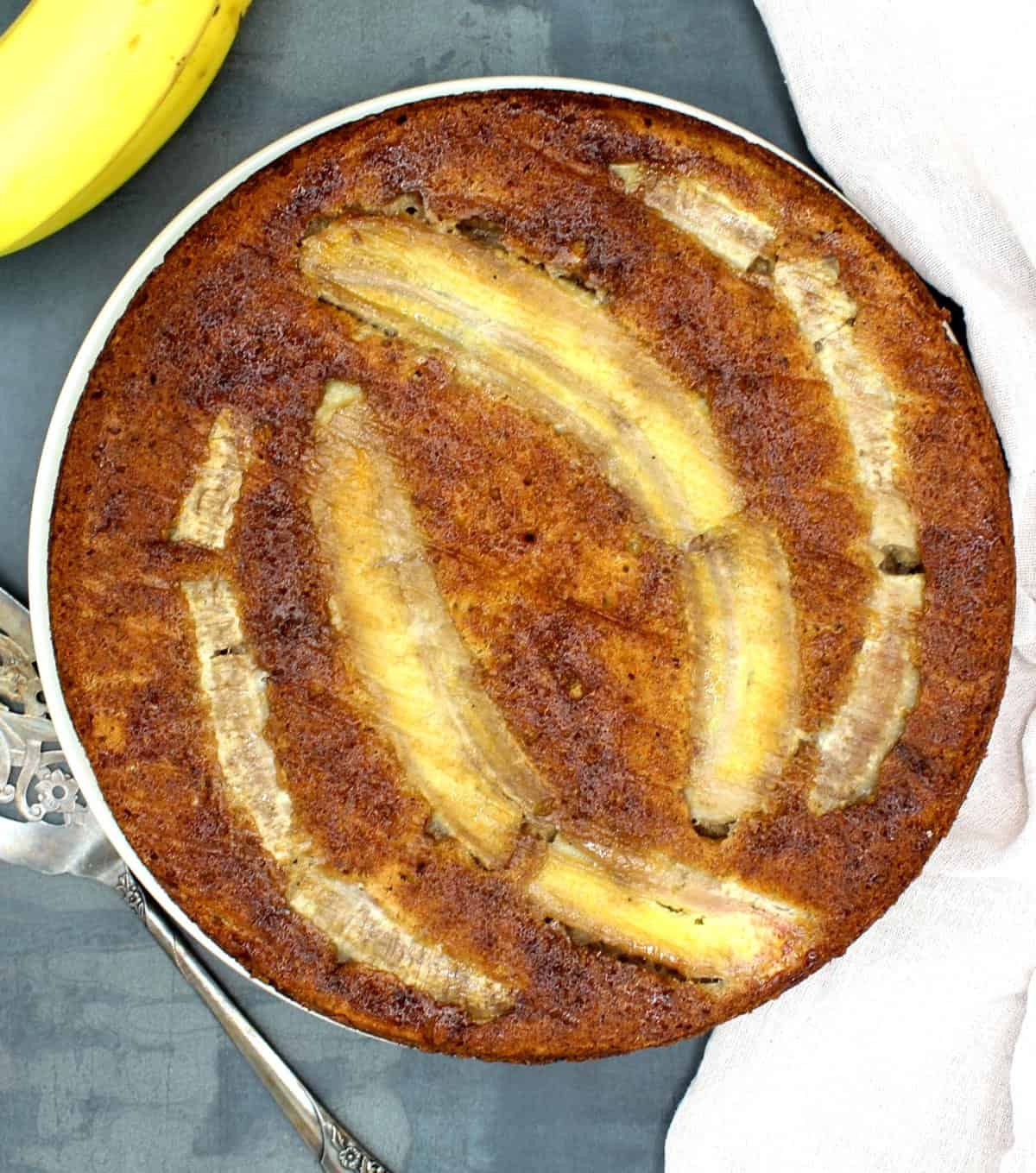 Vegan banana cake with banana in background, and a cake server.