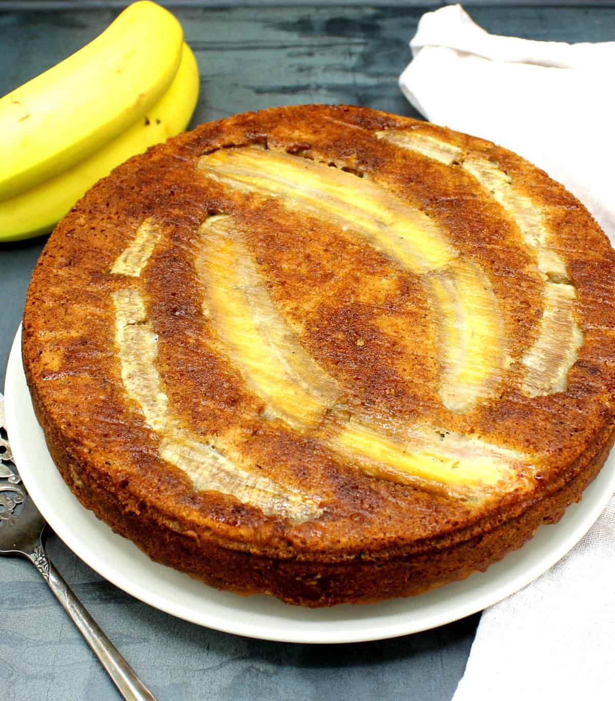 Vegan banana cake on white plate with bananas and cake server in background.