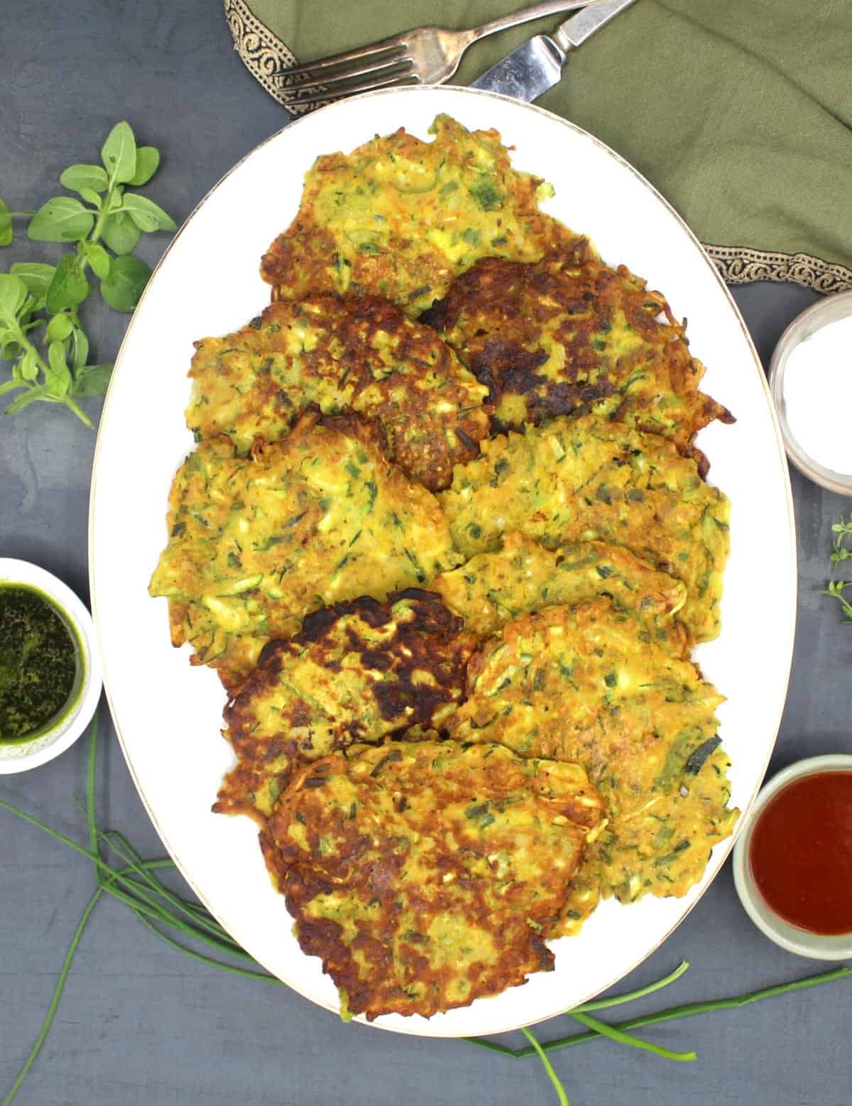 Vegan zucchini fritters on oval white platter surrounded by vegan sauces and herbs and a knife and fork.