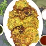 Vegan zucchini fritters in oval plate with sauces and herbs surrounding it.