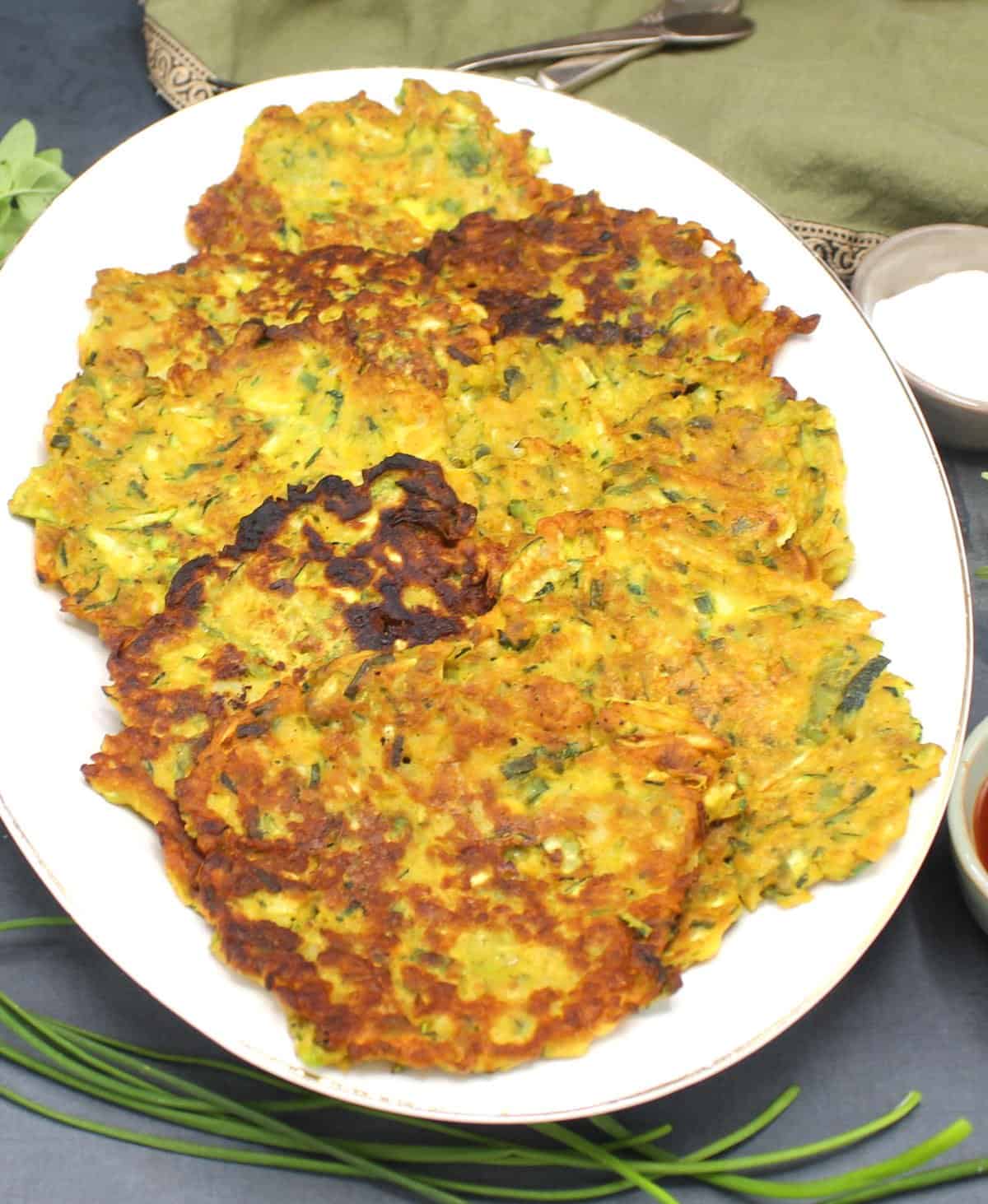 Vegan courgette fritters in white platter with herbs and sauces surrounding it.