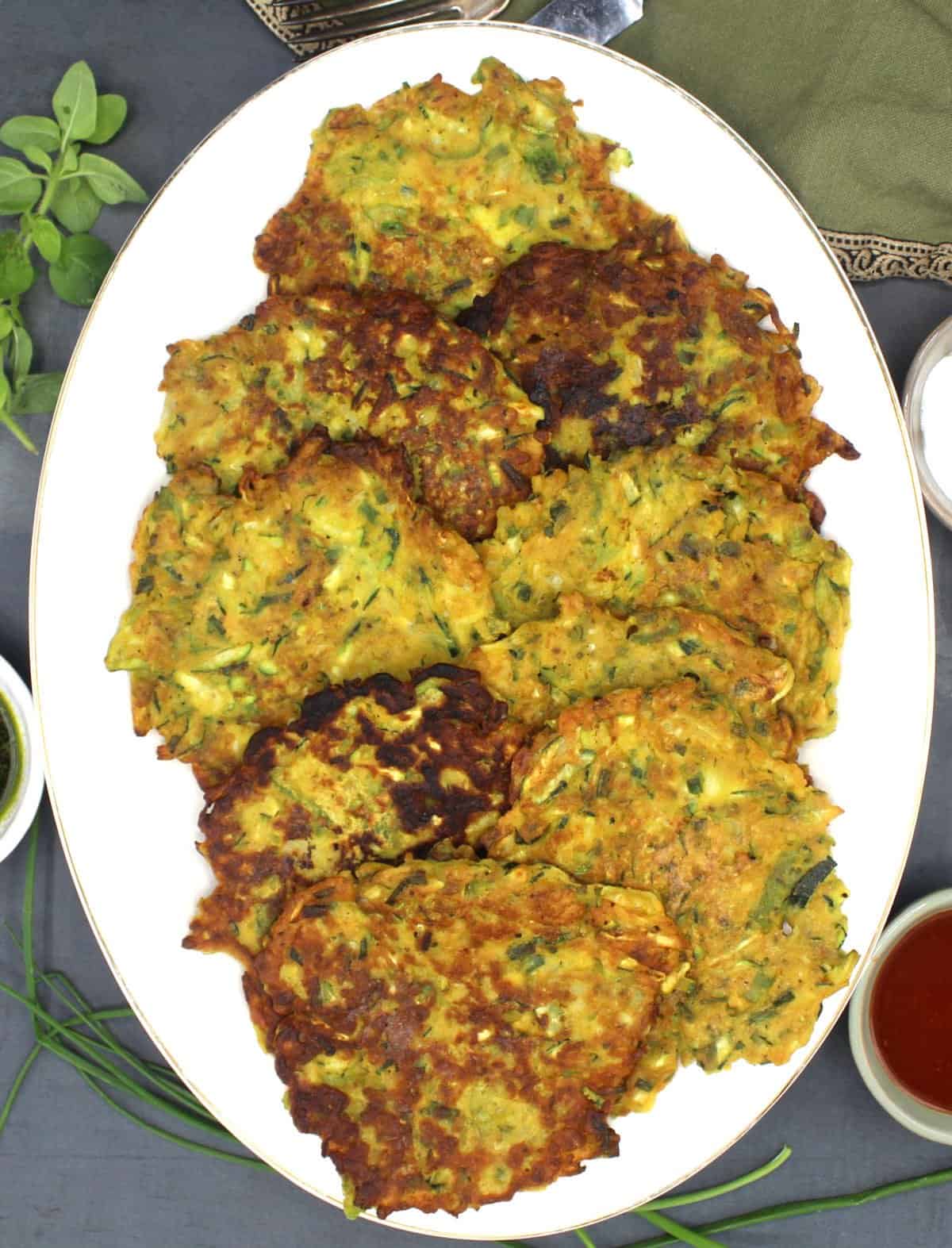 Vegan zucchini fritters on oval white platter with fork, knife, vegan sauces and herbs surrounding it.