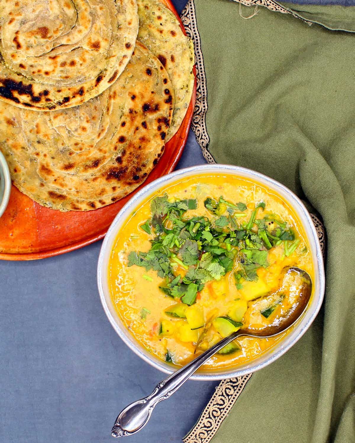 Zucchini dal in bowl with a spoon, cilantro garnish and parathas in background.