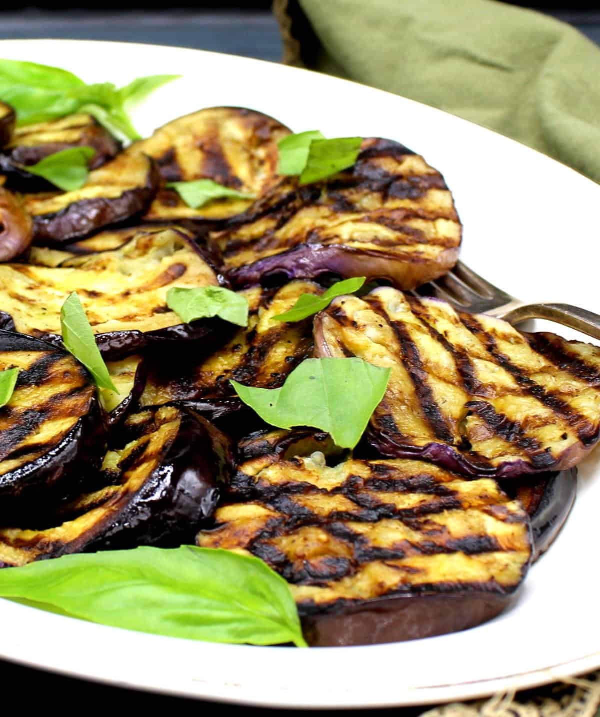 Grilled eggplant slices with basil and a fork in white platter.