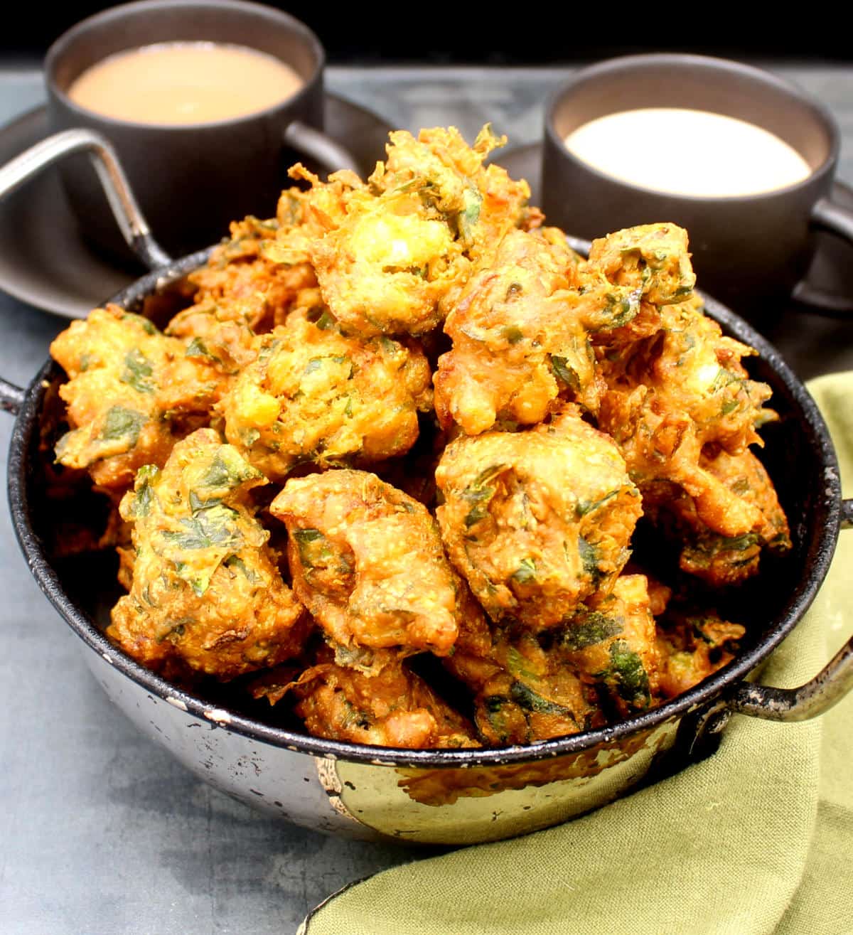 Golden pakora fritters in karahi bowl with cups of chai in background.