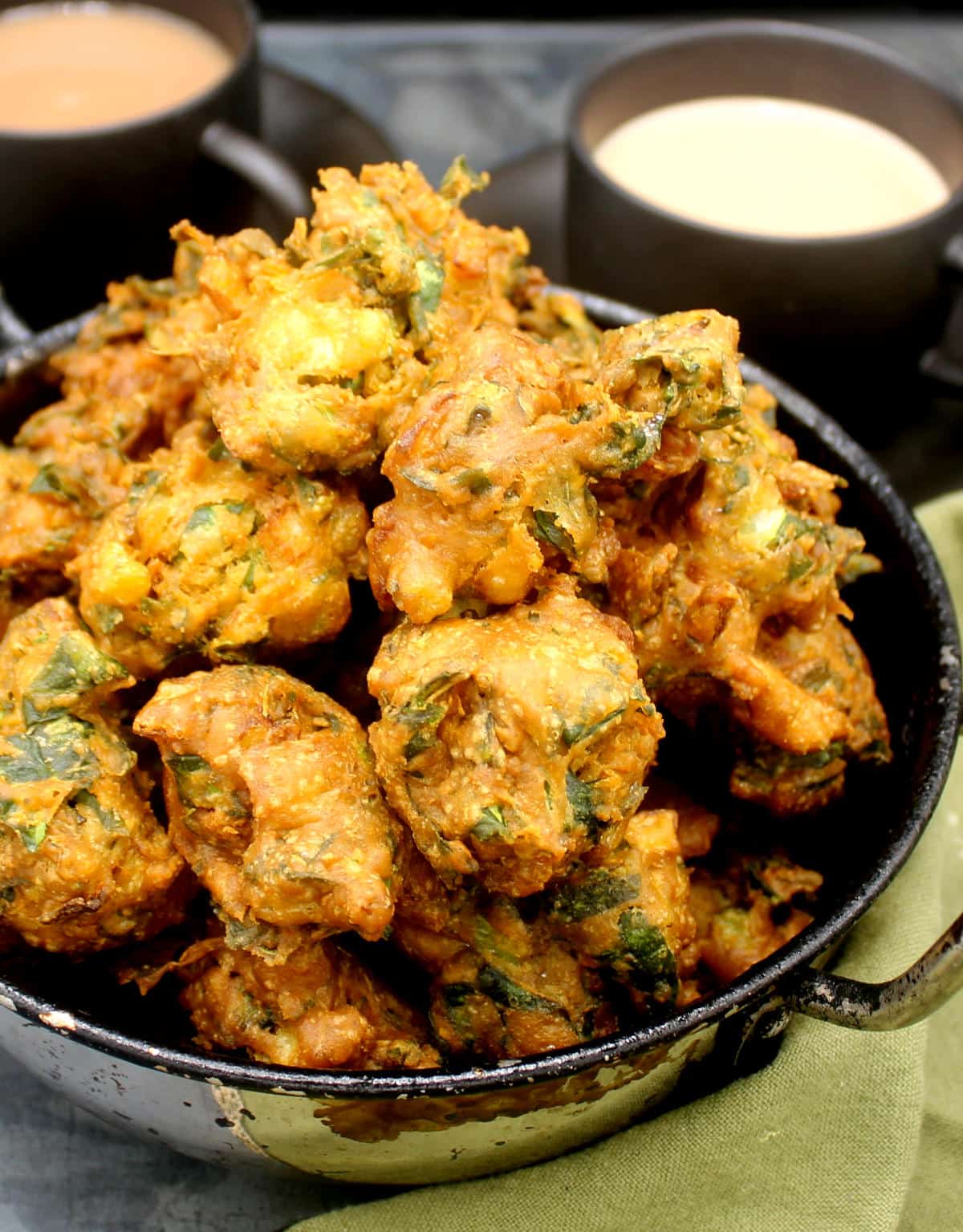 Golden, crunchy pakora fritters made with Swiss chard and onions piled in a karahi bowl with tea in background.