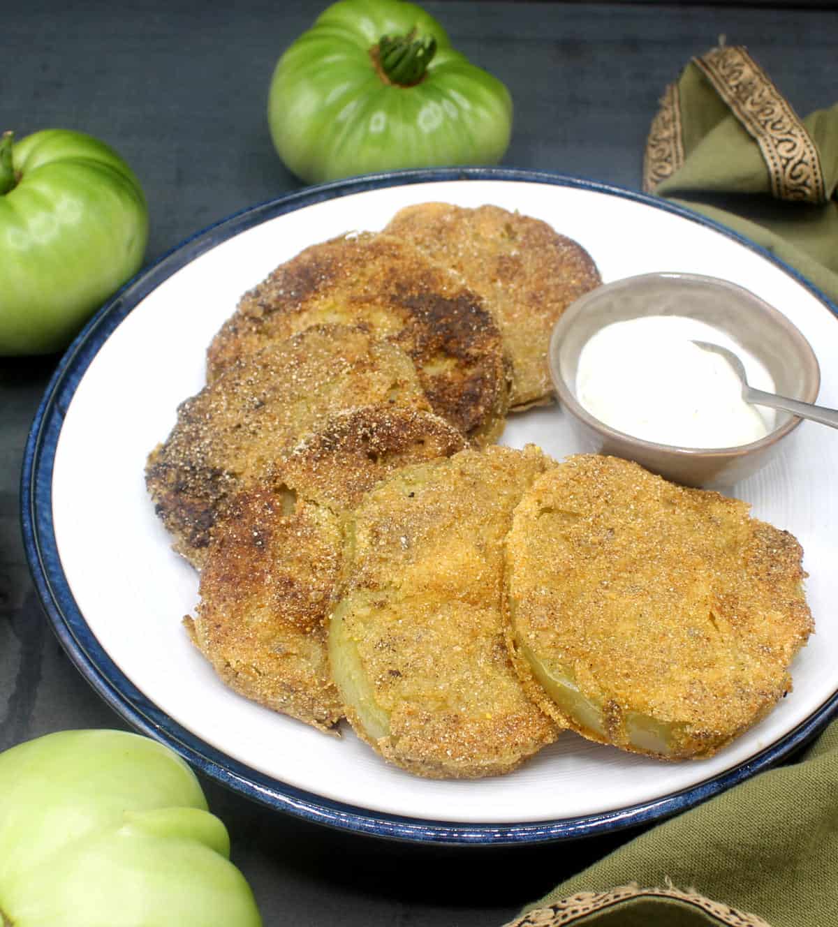 Vegan fried green tomatoes on white plate with spiced mayo in bowl and green tomatoes around it.
