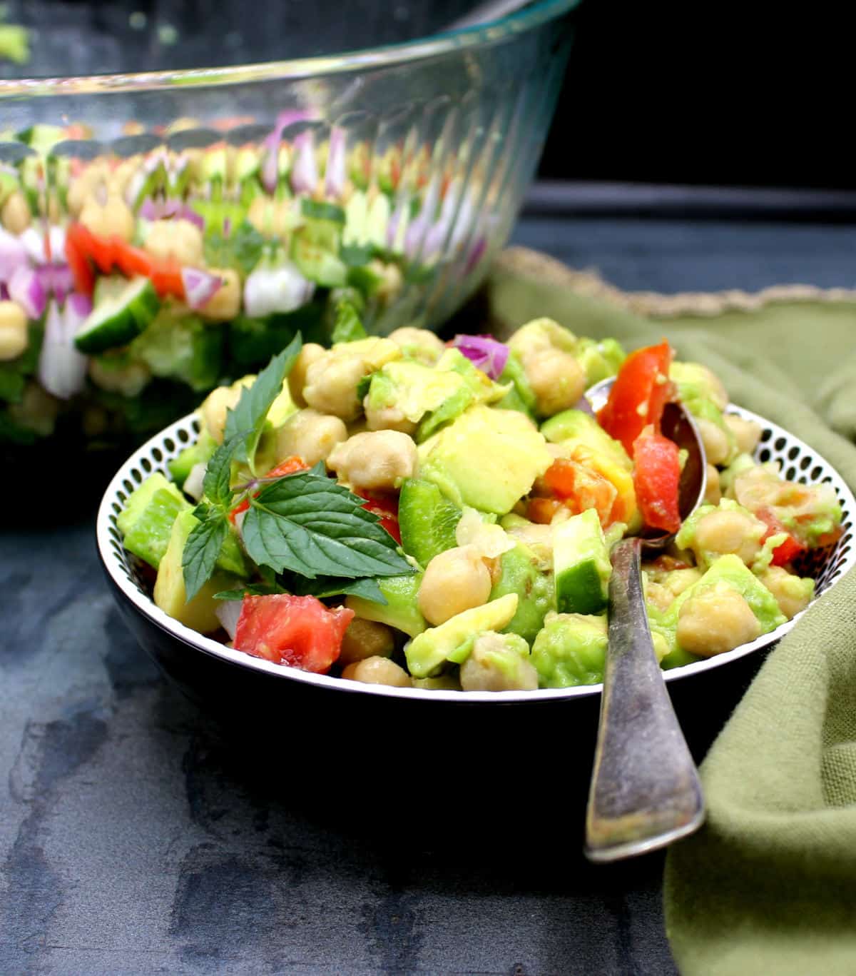 A bowl of chickpea avocado salad with a spoon and big glass salad bowl in background.