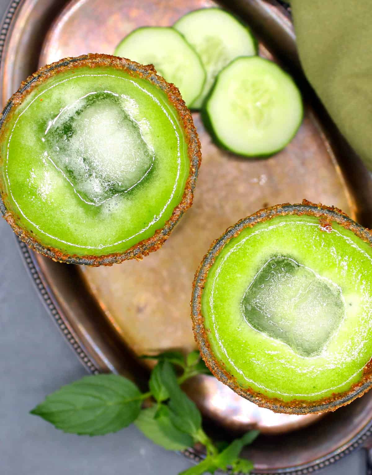 Two glasses of green detoxing cucumber juice with ice. Slices of cucumber and mint are next to the glasses, on a silver tray.