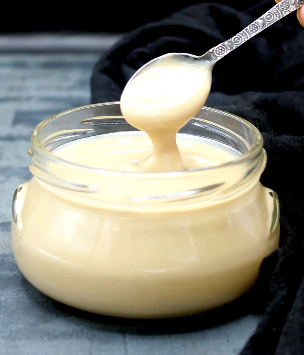 Creamy vegan condensed milk being poured into glass jar with decorative silver spoon. 