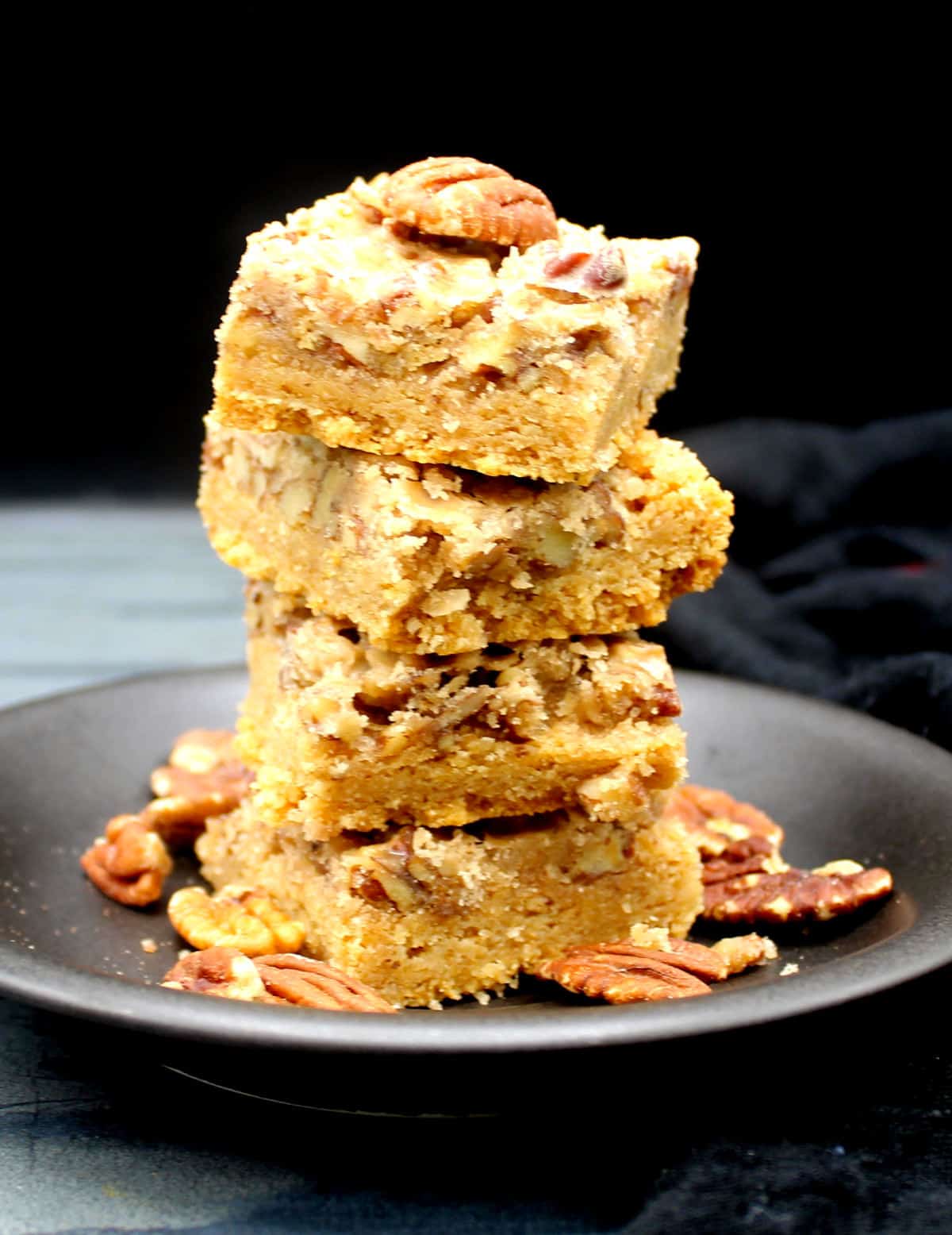 A stack of vegan pecan squares in black plate with walnuts scattered around.