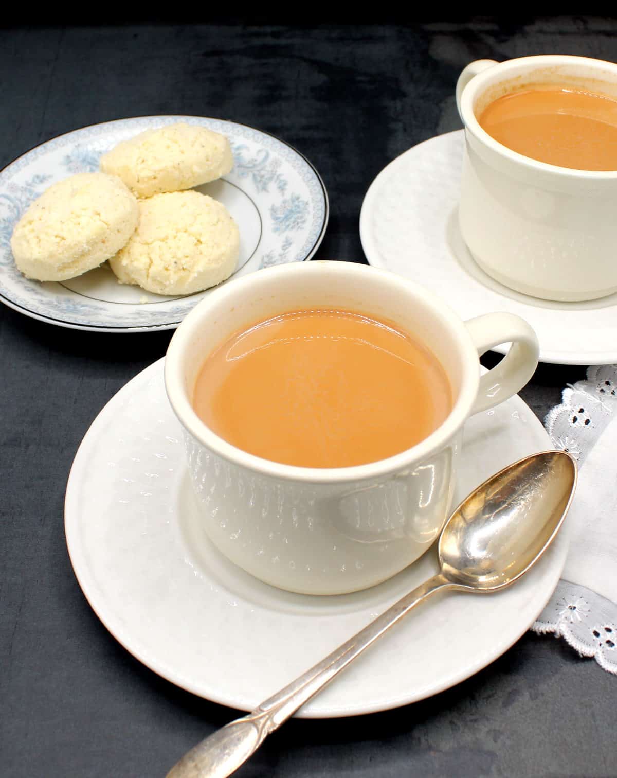 Masala chai tea in white cups and saucers with a plate of nankhatai biscuits in background.