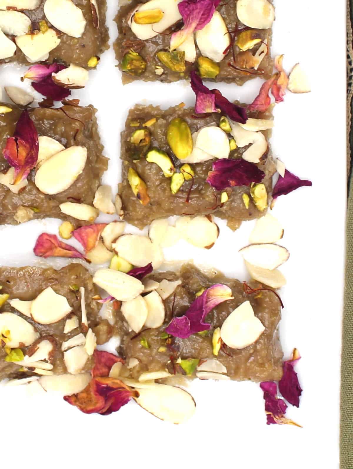 Rectangle vegan kalakand pieces on a plate with rose petals, nuts and saffron strands.