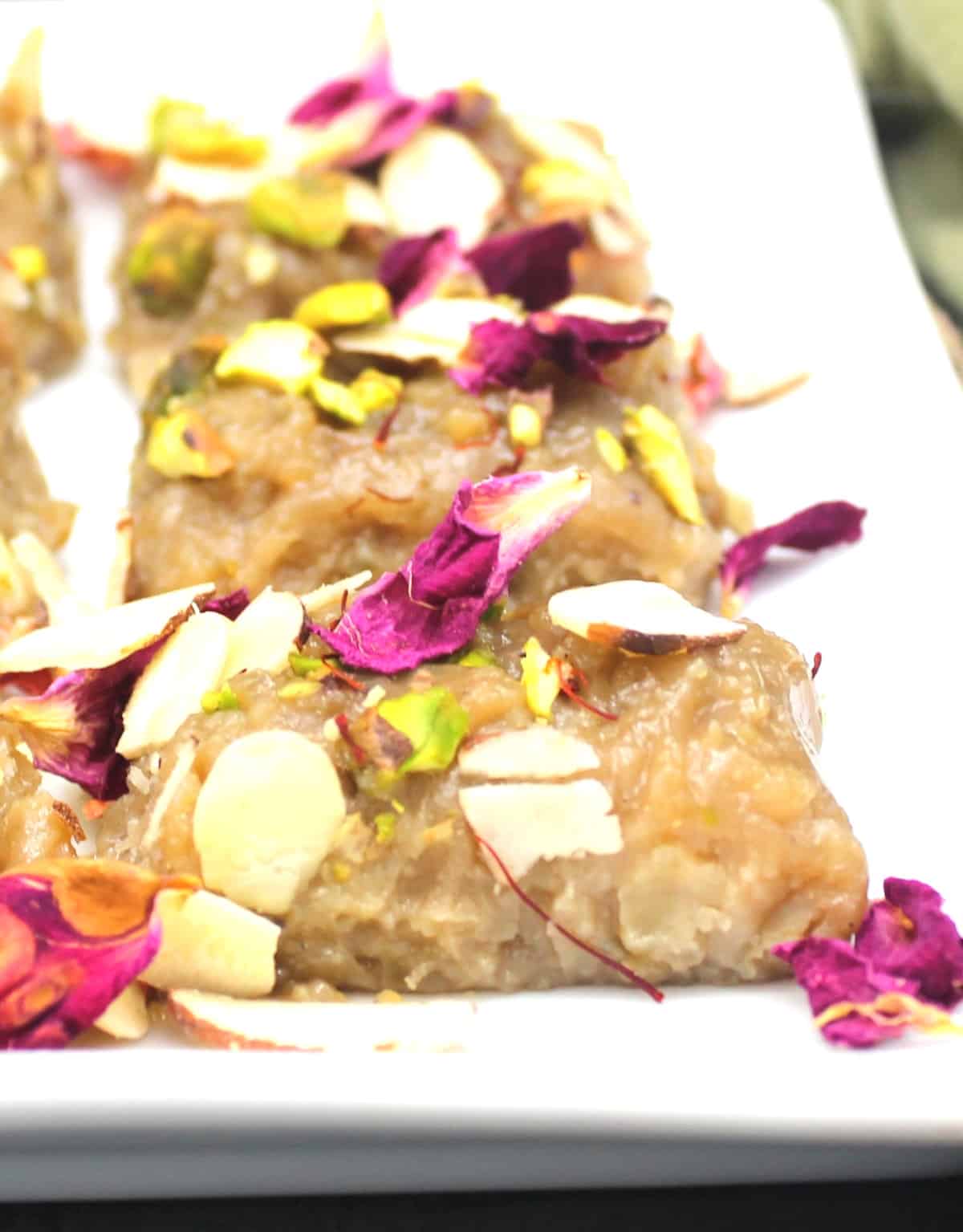Vegan kalakand in white plate with rose petals, nuts and saffron garnish.