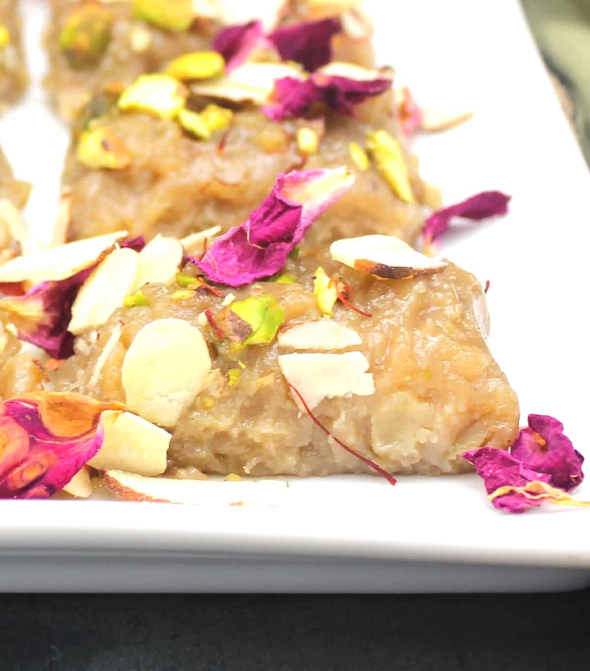 Vegan kalakand pieces on white plate with rose petals and nuts.