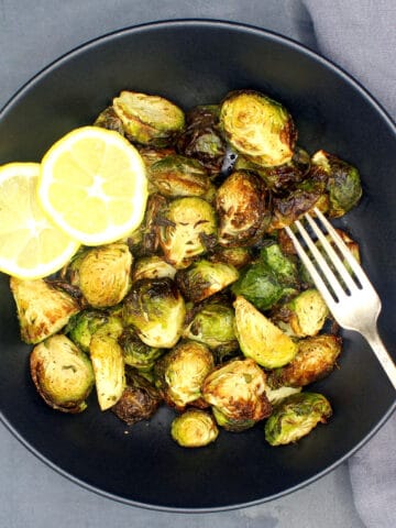 Roasted air fried Brussels sprouts in black bowl with slices of lemon and fork.