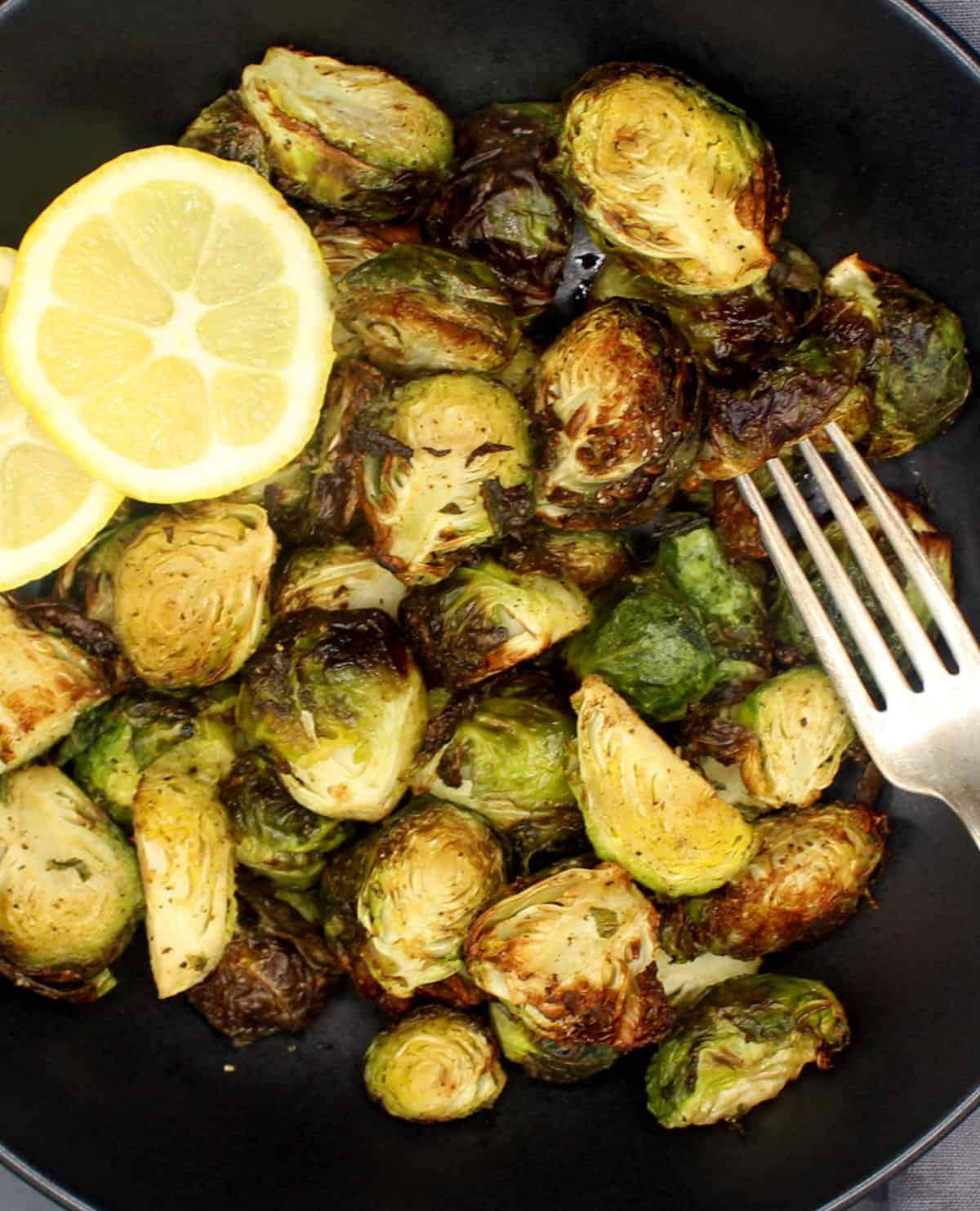 Air fryer Brussels sprouts with slices of lemon and a fork in black bowl.
