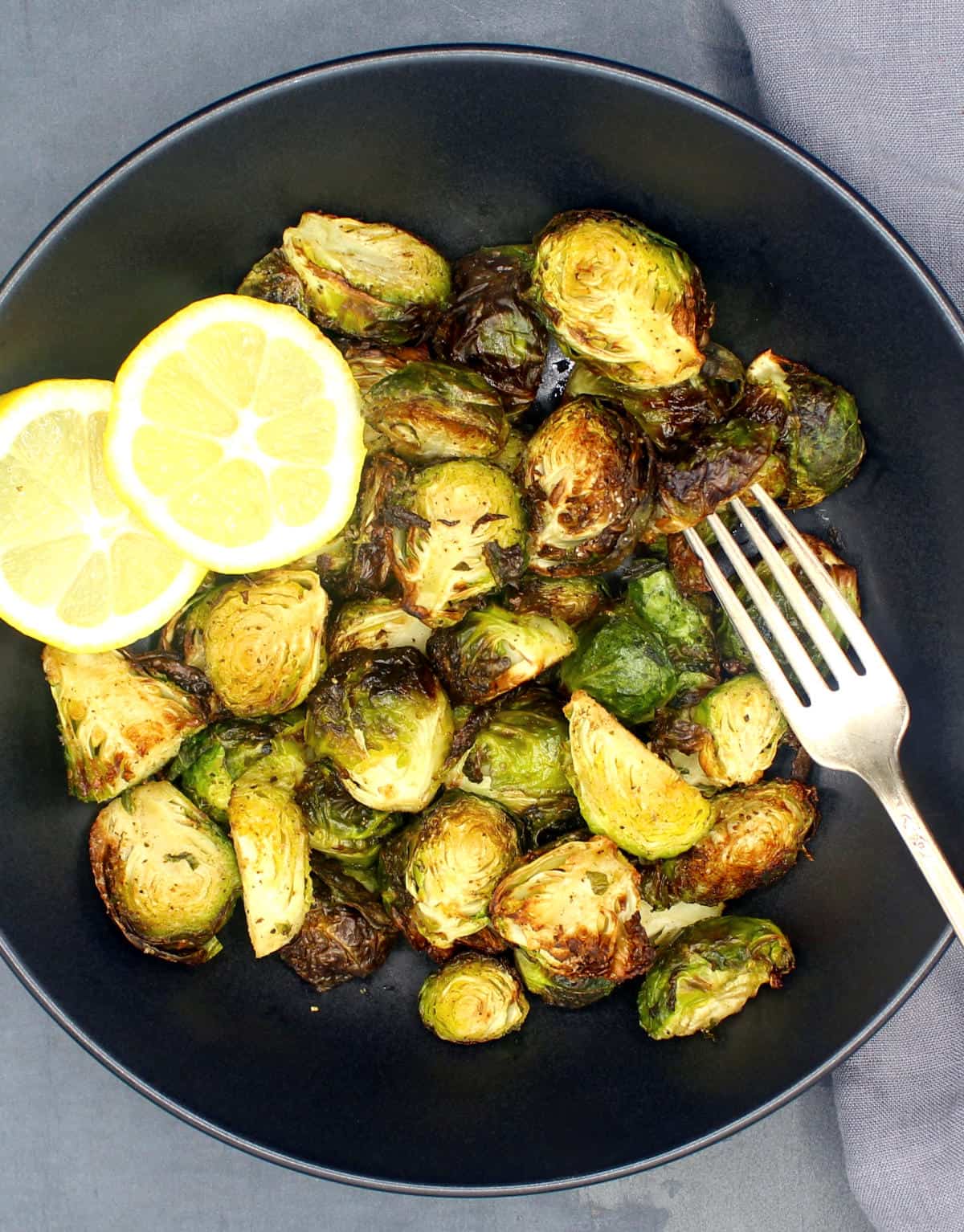 Roasted air fried Brussels sprouts in black bowl with slices of lemon and fork.