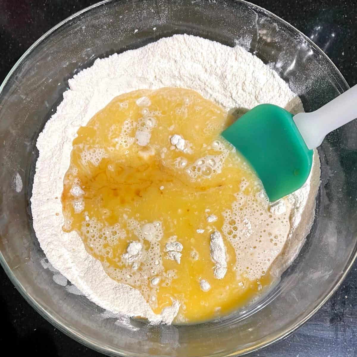 Wet ingredients added to dry in glass bowl with spatula.