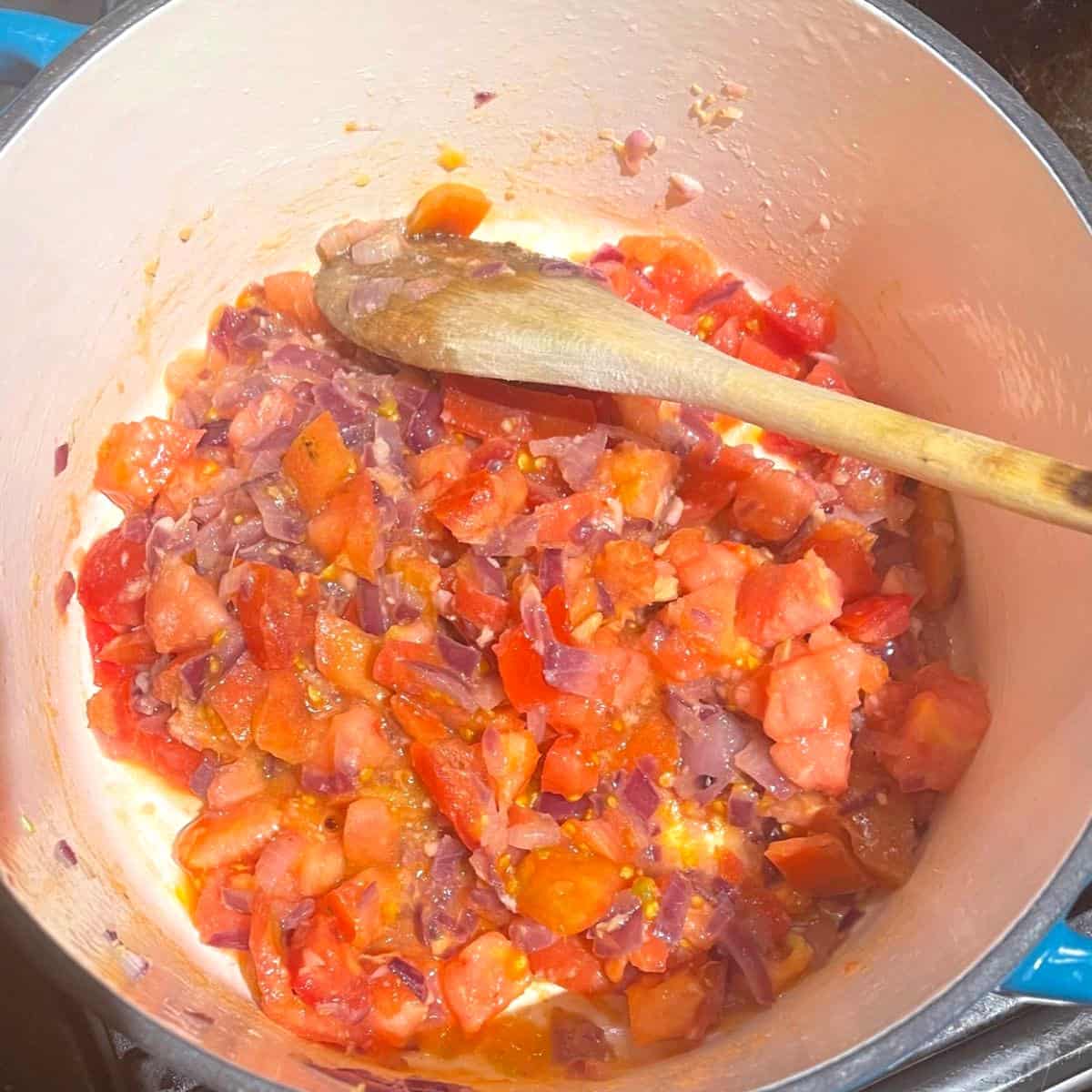 Tomatoes and onions sauteing in pot with wooden spatula.