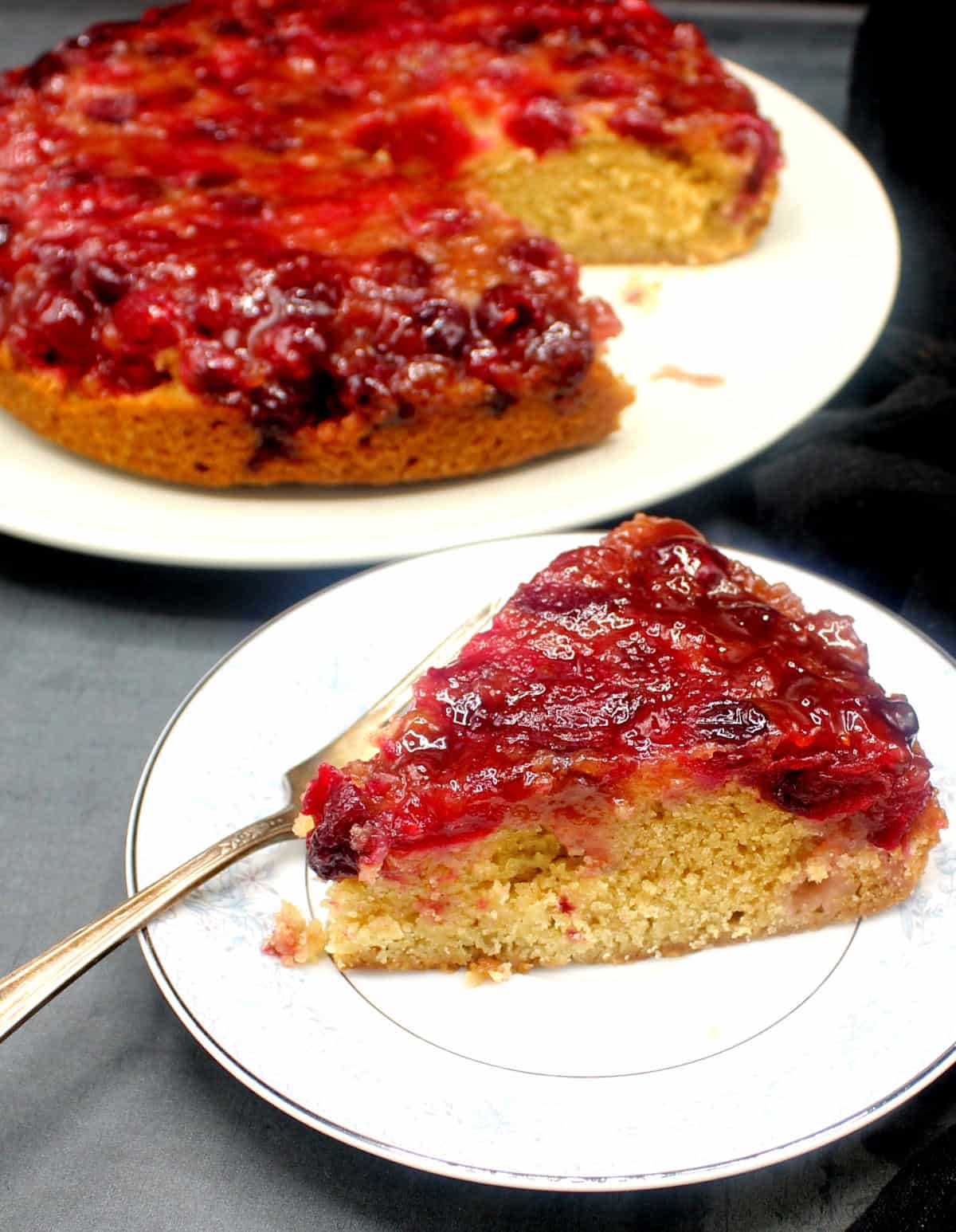 Slice of vegan cranberry upside down cake in plate with full cake in background.