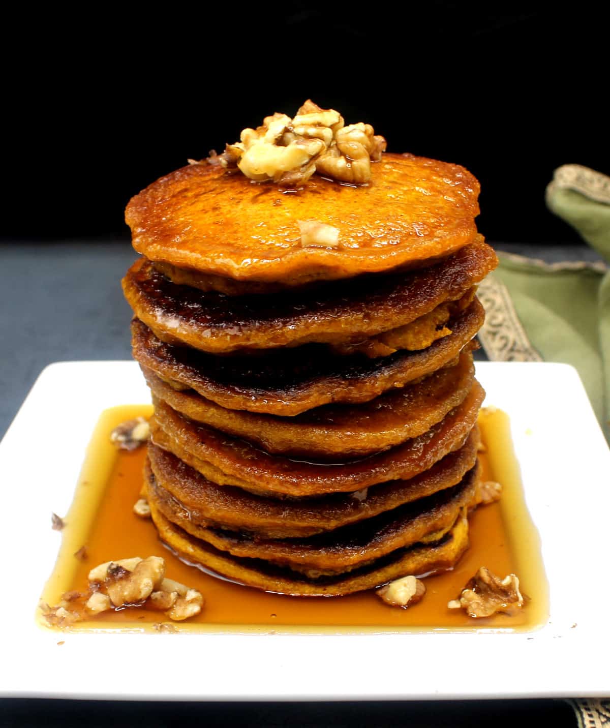 A stack of vegan pumpkin pancakes topped with walnuts in a pool of maple syrup.