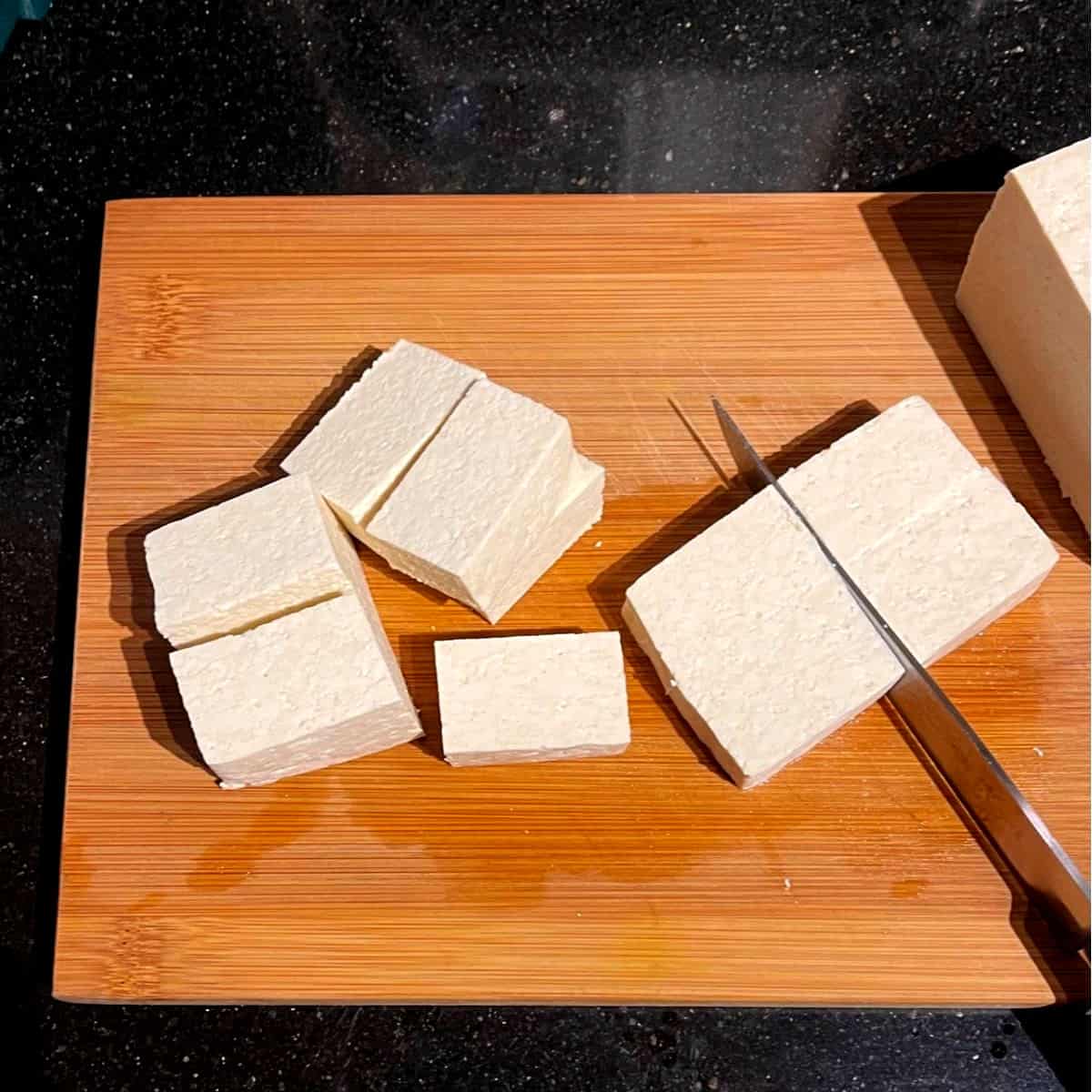 Tofu cut in slices on chopping board with knife.