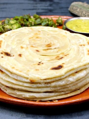 Parathas stacked on a clay plate with dal and sabzi.