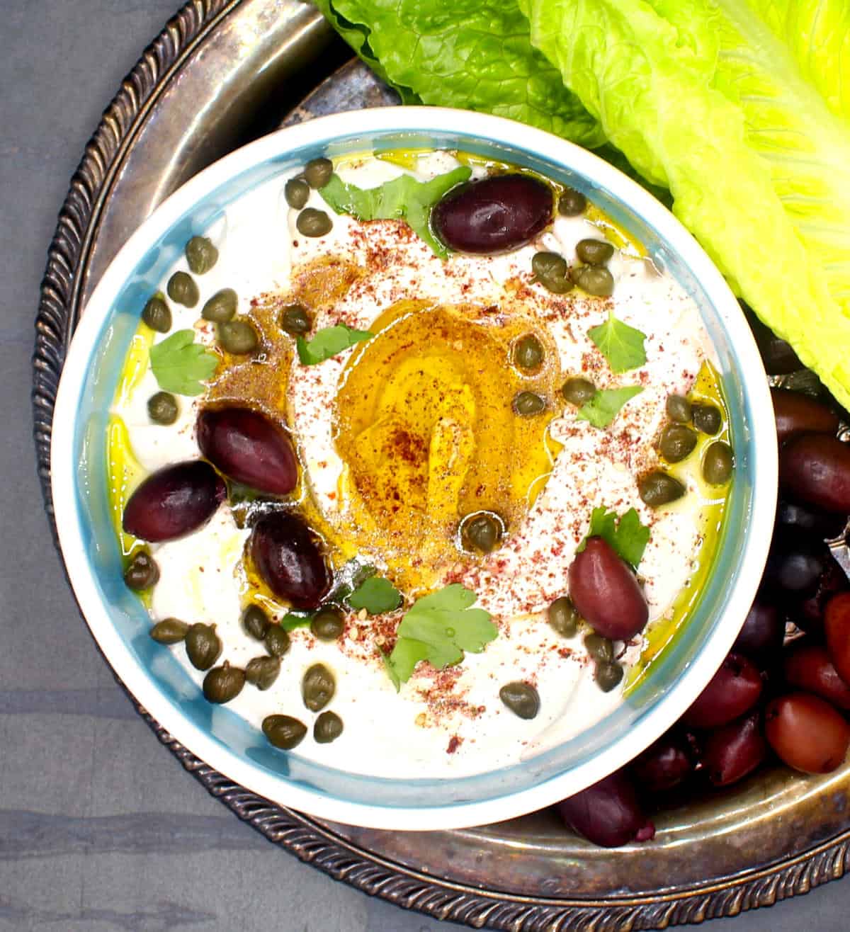 Vegan labneh with za'atar, olive oil, capers and parsley in a bowl with lettuce and olives on the side.