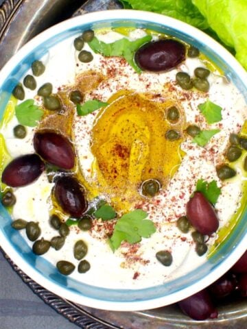 Vegan labneh with za'atar, olive oil, capers and parsley in bowl with lettuce and olives on the side.