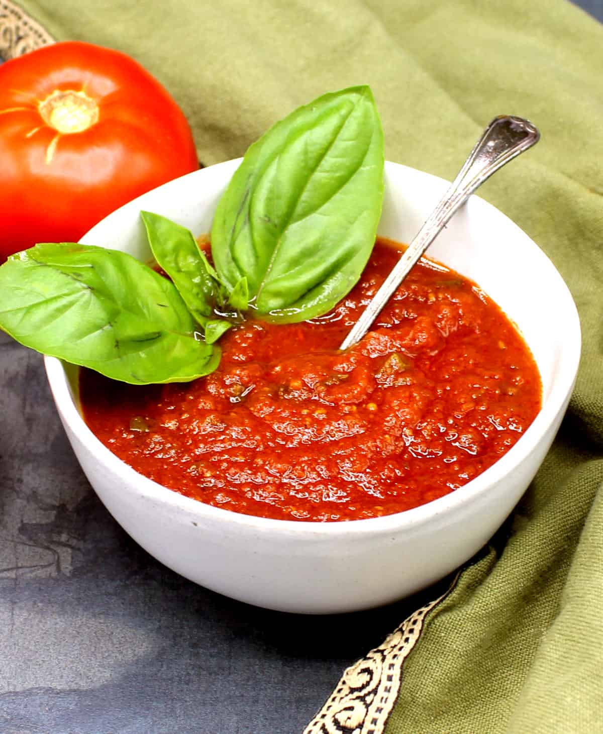 Homemade tomato sauce in white bowl with basil leaves. In the background is a tomato and a green napkin.