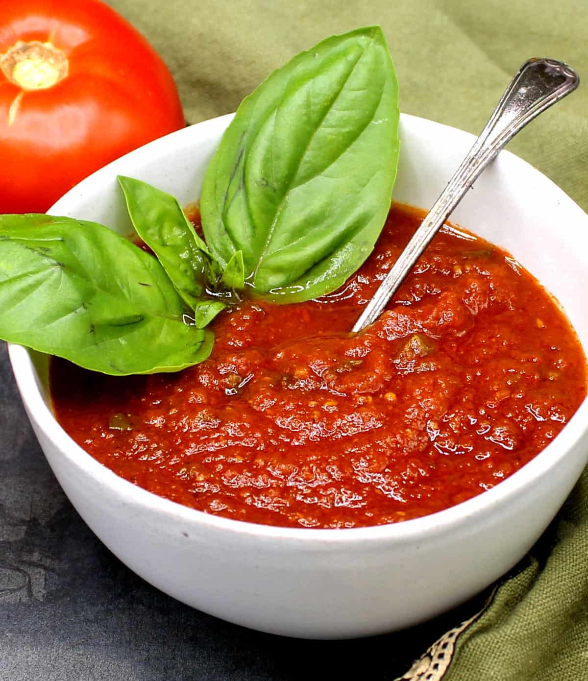 Homemade tomato sauce in bowl with a sprig of basil and a fresh tomato in background.