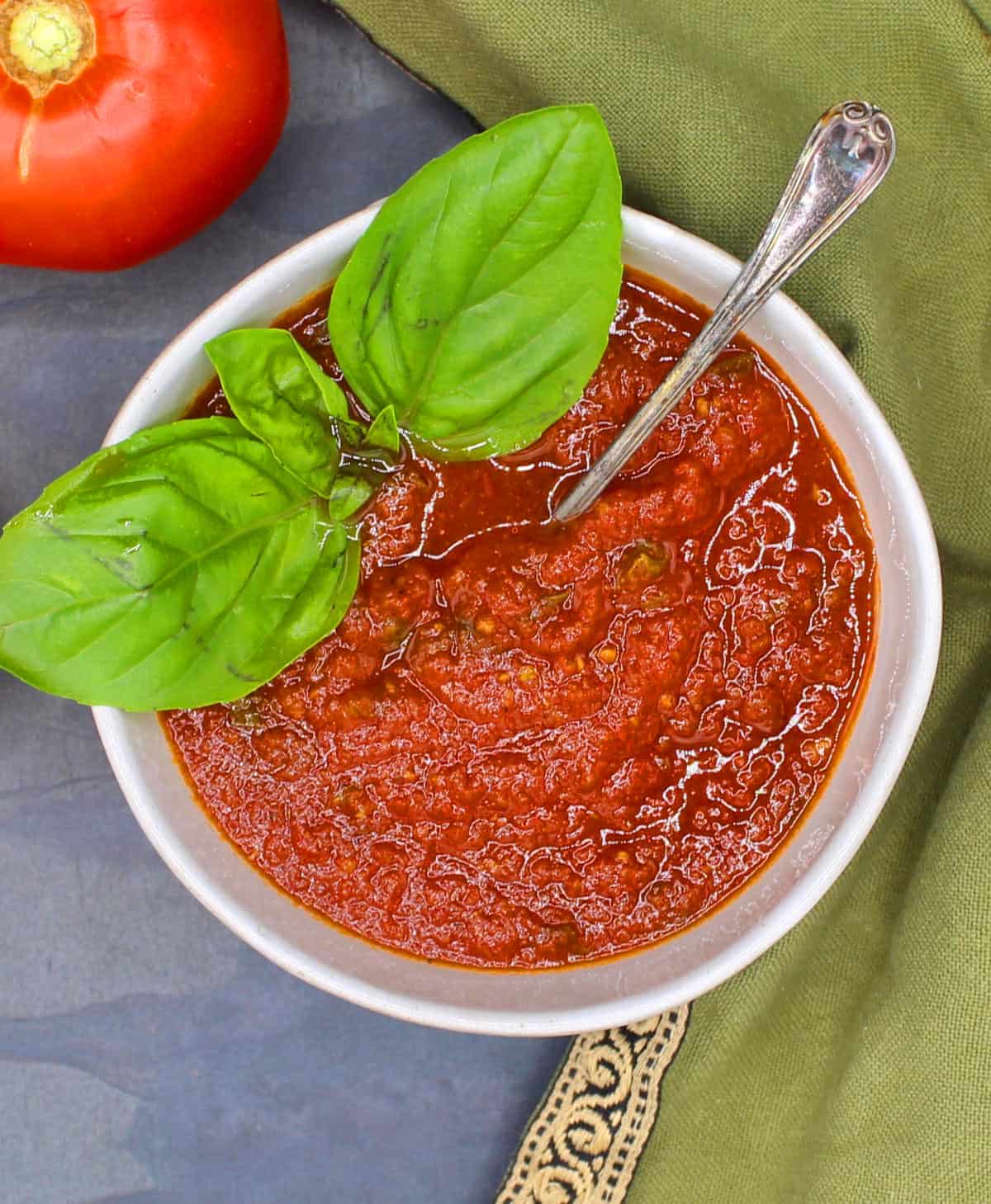 Tomato sauce in bowl with basil leaves and tomato in background.