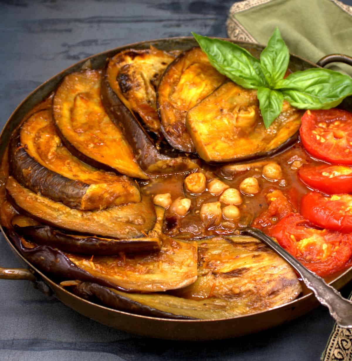 Khoresh bademjan in a copper pot with eggplant slices, tomatoes and basil garnish.