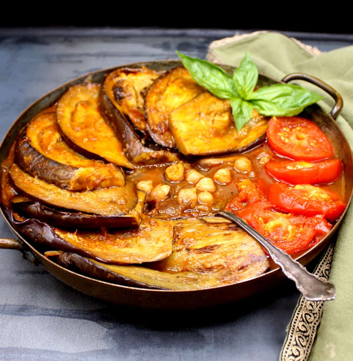 Khoresh bademjan in a copper pot with eggplant slices, tomatoes and basil garnish.