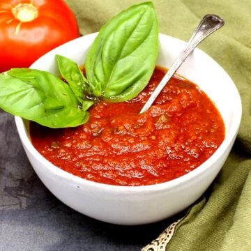 Sos pomidorowy w misce z łyżką i liśćmi świeżej bazylii oraz pomidorem w tle.
