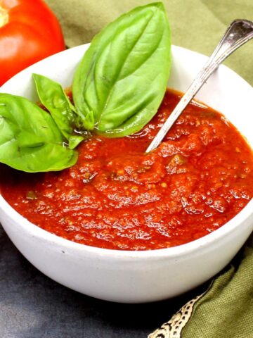 Tomato sauce in bowl with fresh basil leaves and spoon and a tomato in background.