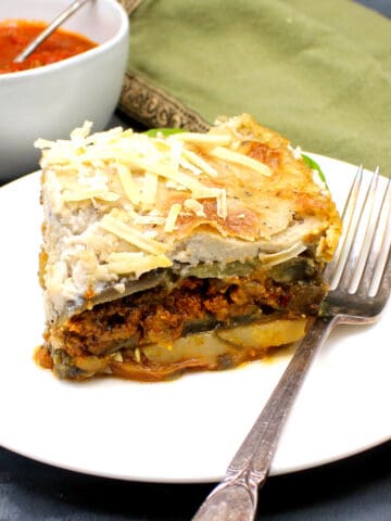 A slice of vegan moussaka with layers of potato, zucchini, eggplant, vegan beef ragu and a creamy vegan bechamel sauce, in white plate with fork. Tomato sauce in background.